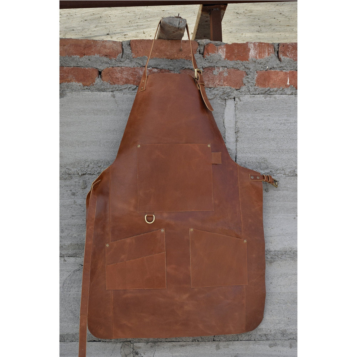 May include: A brown leather apron with multiple pockets and a brass buckle. The apron is hanging on a brick wall.