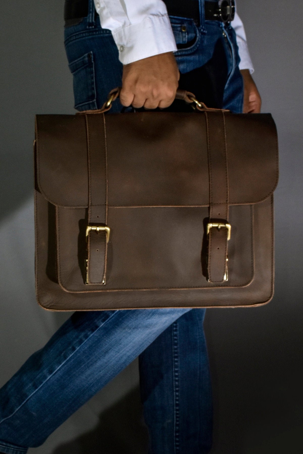 May include: A brown leather briefcase with two brass buckles and a top handle. The briefcase is being held by a person wearing blue jeans.