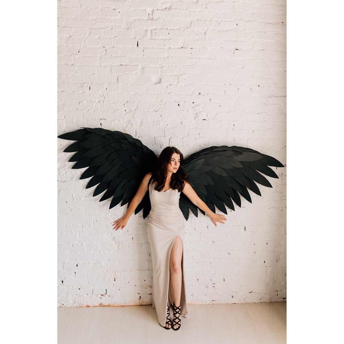 May include: A woman wearing a beige dress and black strappy heels stands in front of a white brick wall, with large black paper angel wings attached to the wall behind her. The dress has a high slit.