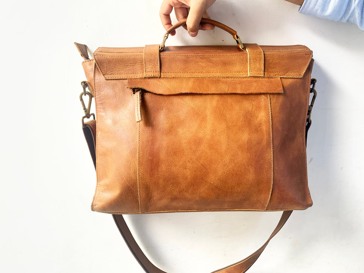 May include: A brown leather messenger bag with a top handle and adjustable shoulder strap. The bag has a flap closure with a buckle and a zippered pocket on the front.