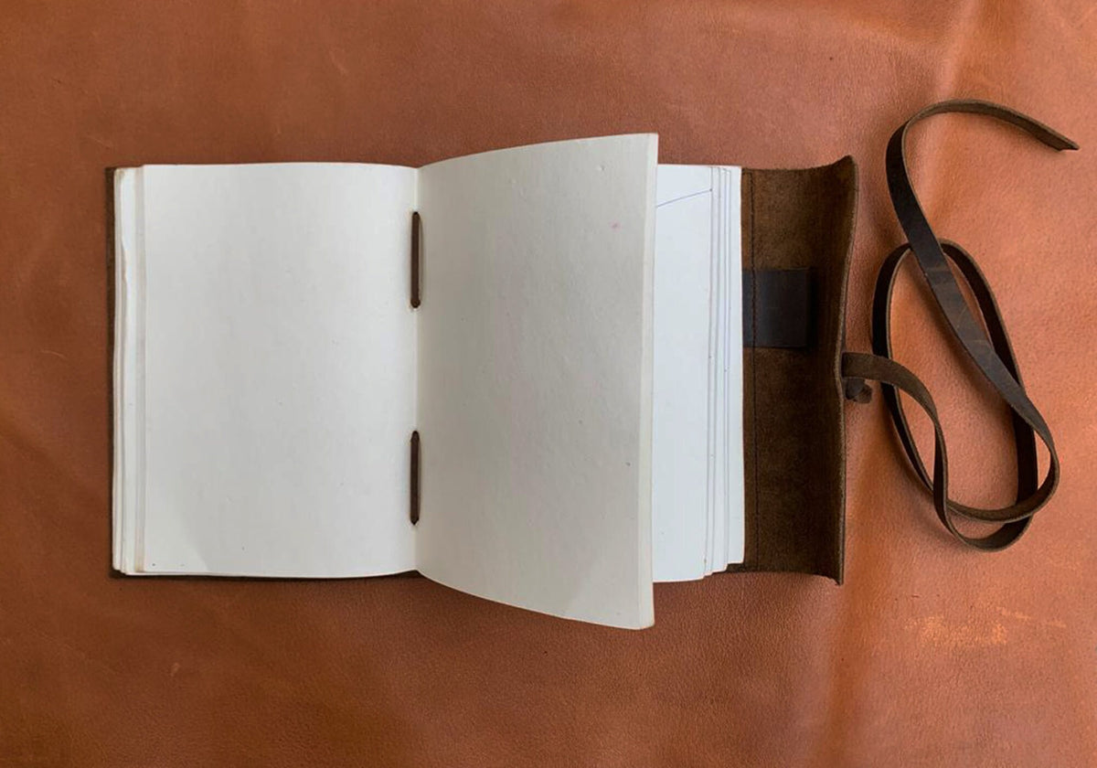 May include: A brown leather-bound journal with blank pages, open to reveal the white paper inside. The journal has a brown leather strap and a brown leather closure.