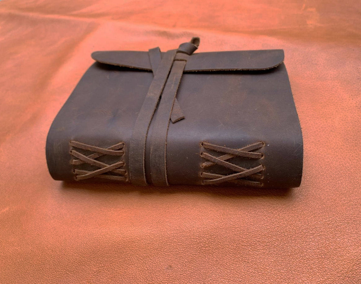 May include: A brown leather journal with a flap closure and a leather strap. The journal has a decorative stitching pattern on the spine.