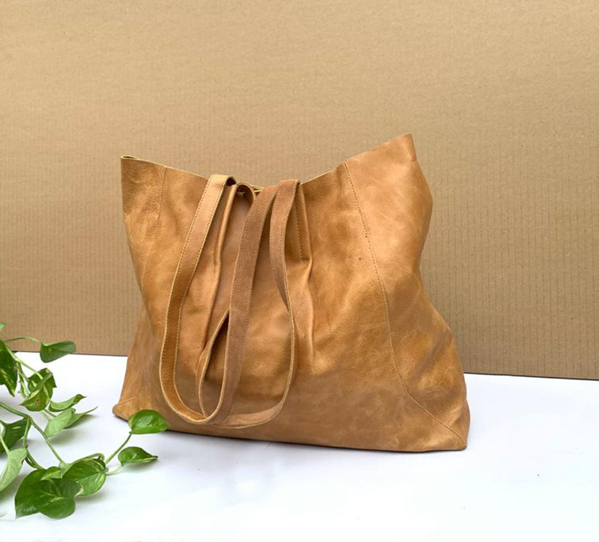 May include: A large, tan leather tote bag with long straps. The bag has a soft, worn appearance and is made of a single piece of leather. The straps are a lighter shade of brown. The bag is sitting on a white surface.