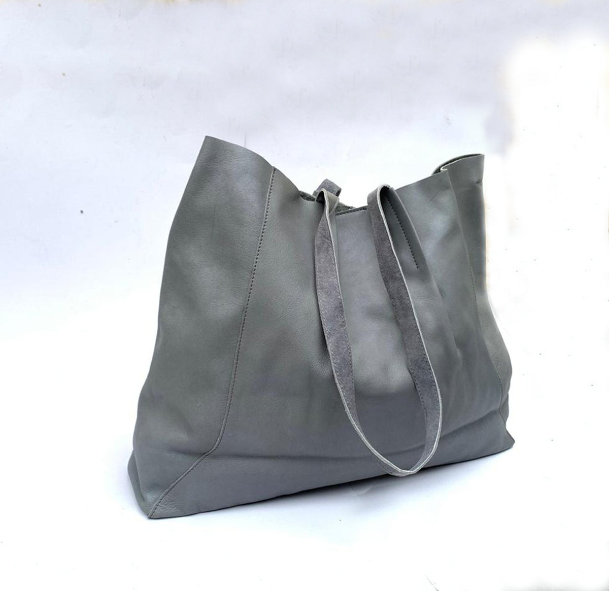 May include: A large, gray leather tote bag with two long straps. The bag is made of a soft, supple leather and has a simple, minimalist design. The bag is spacious and appears to be empty. The background is white.