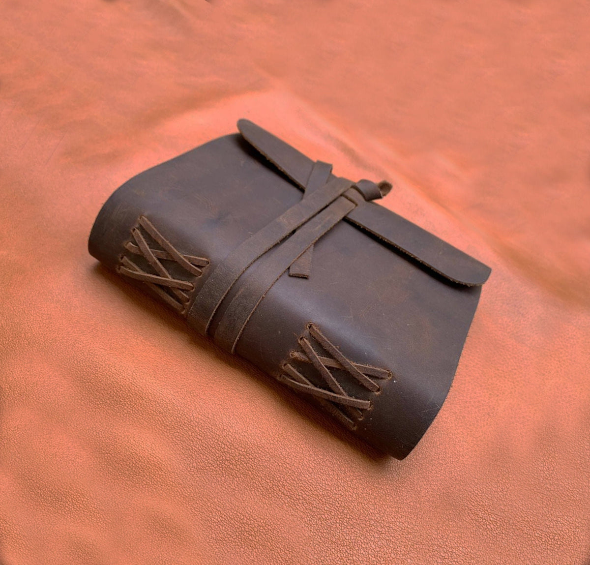 May include: A brown leather journal with a strap closure and decorative stitching. The journal is closed and lying on a brown leather surface.