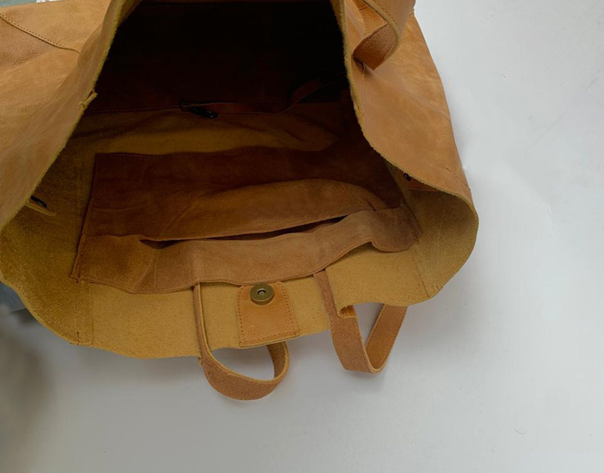 May include: A large, open-top tote bag made of tan leather. The bag has two handles and a snap closure. The interior features a large pocket and a smaller pocket. The leather has a natural, textured appearance.