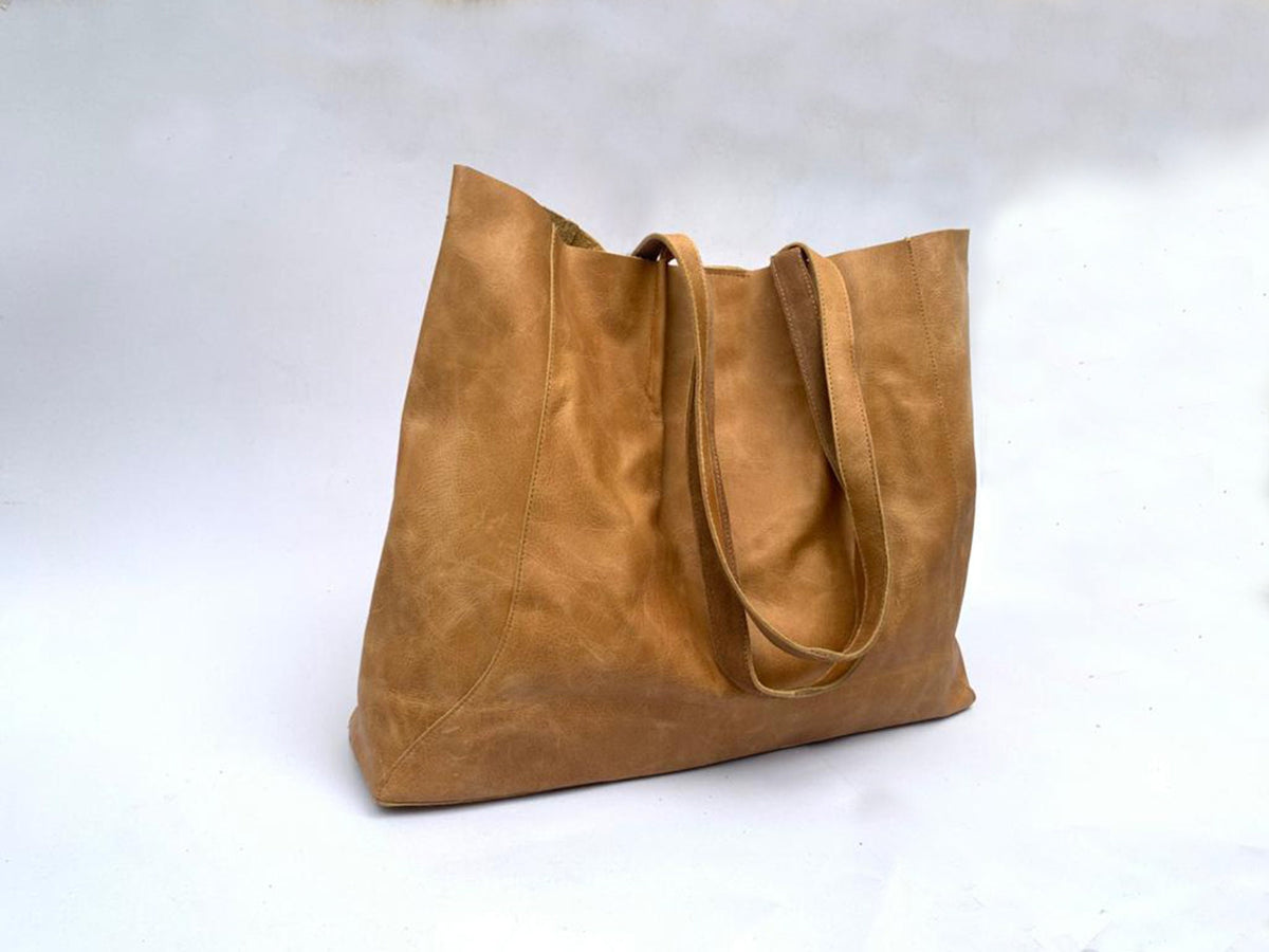 May include: A large, tan leather tote bag with two long handles. The bag has a rectangular shape with a wide opening and a slightly distressed appearance. The leather has a natural, textured look.