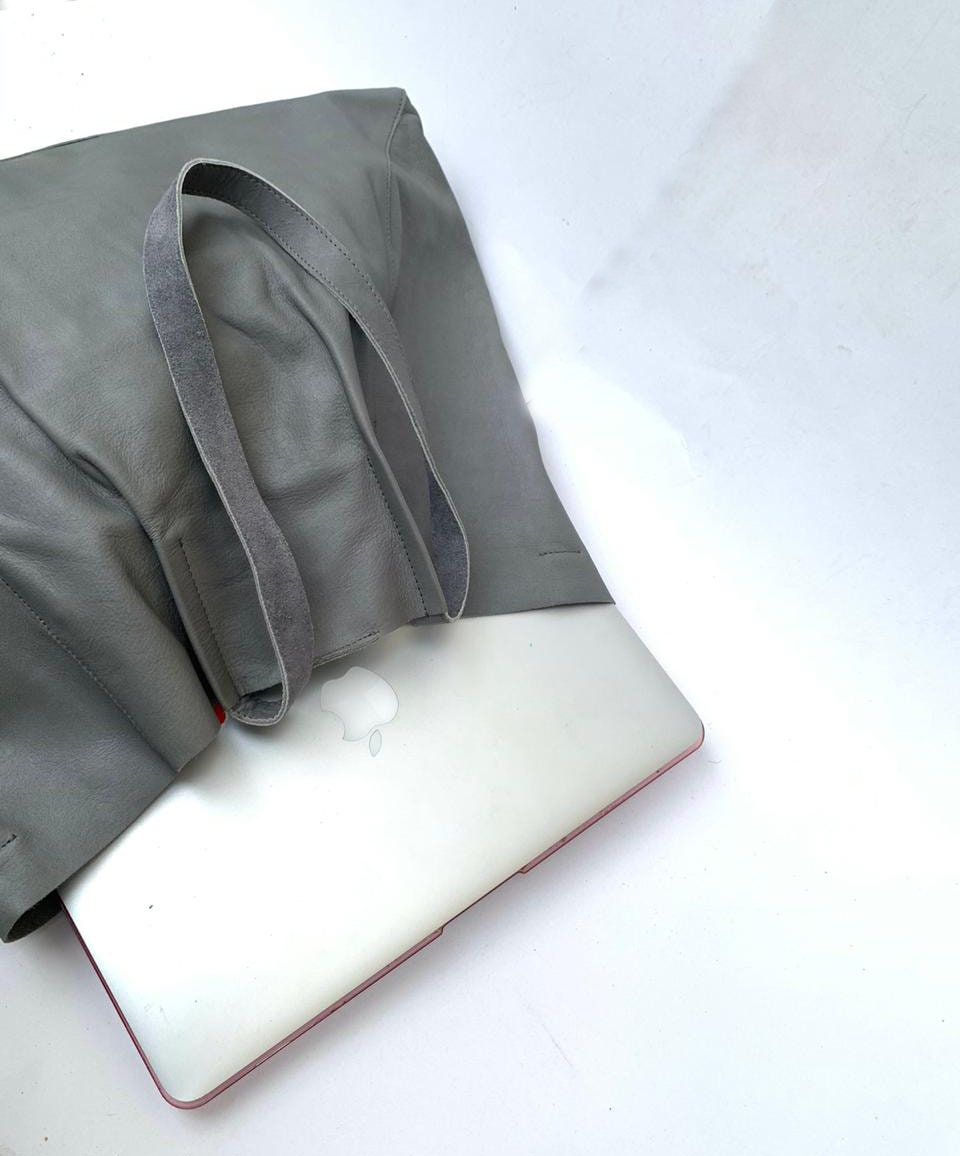 May include: A gray leather tote bag with two handles, holding a silver laptop. The bag is made of soft, textured leather. The laptop has a silver finish and a red border. The bag is designed to carry a laptop and other essentials.