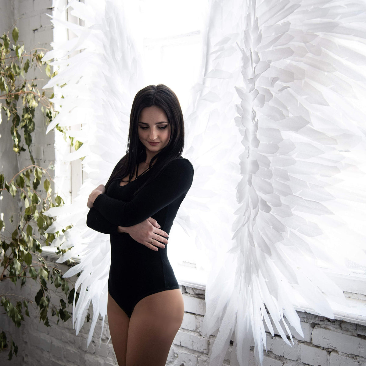 May include: A woman wearing a black bodysuit stands in front of a white backdrop with large white angel wings. The woman is looking down and has her arms crossed.
