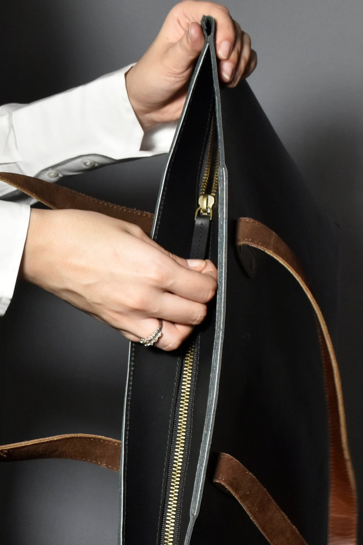 May include: A black leather tote bag with brown leather straps and a gold zipper. The bag is open and the zipper is pulled down.