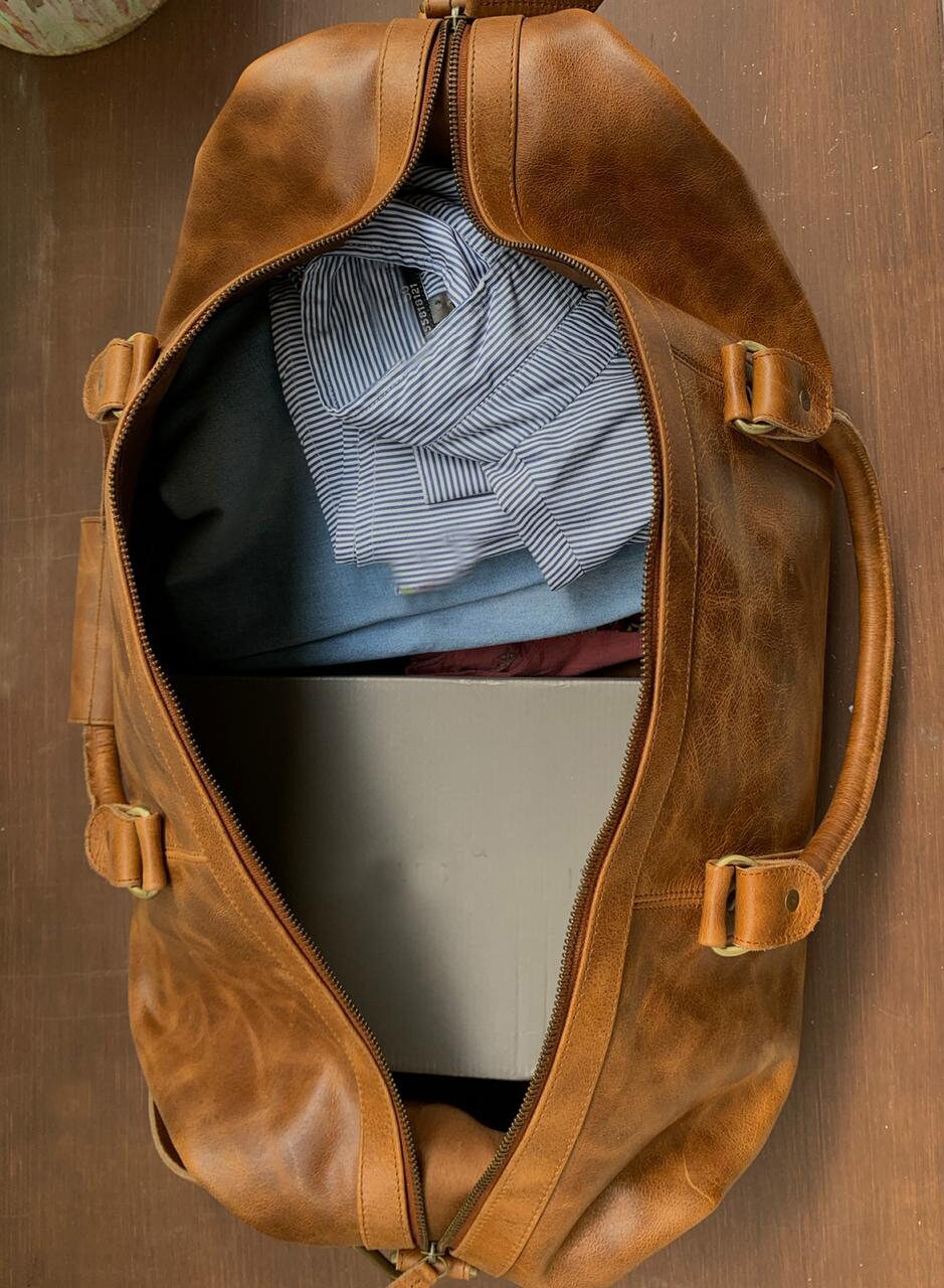 May include: A brown leather duffel bag with a zipper closure and two straps. The bag is open and contains a blue and white striped shirt, a blue shirt, and a white box.