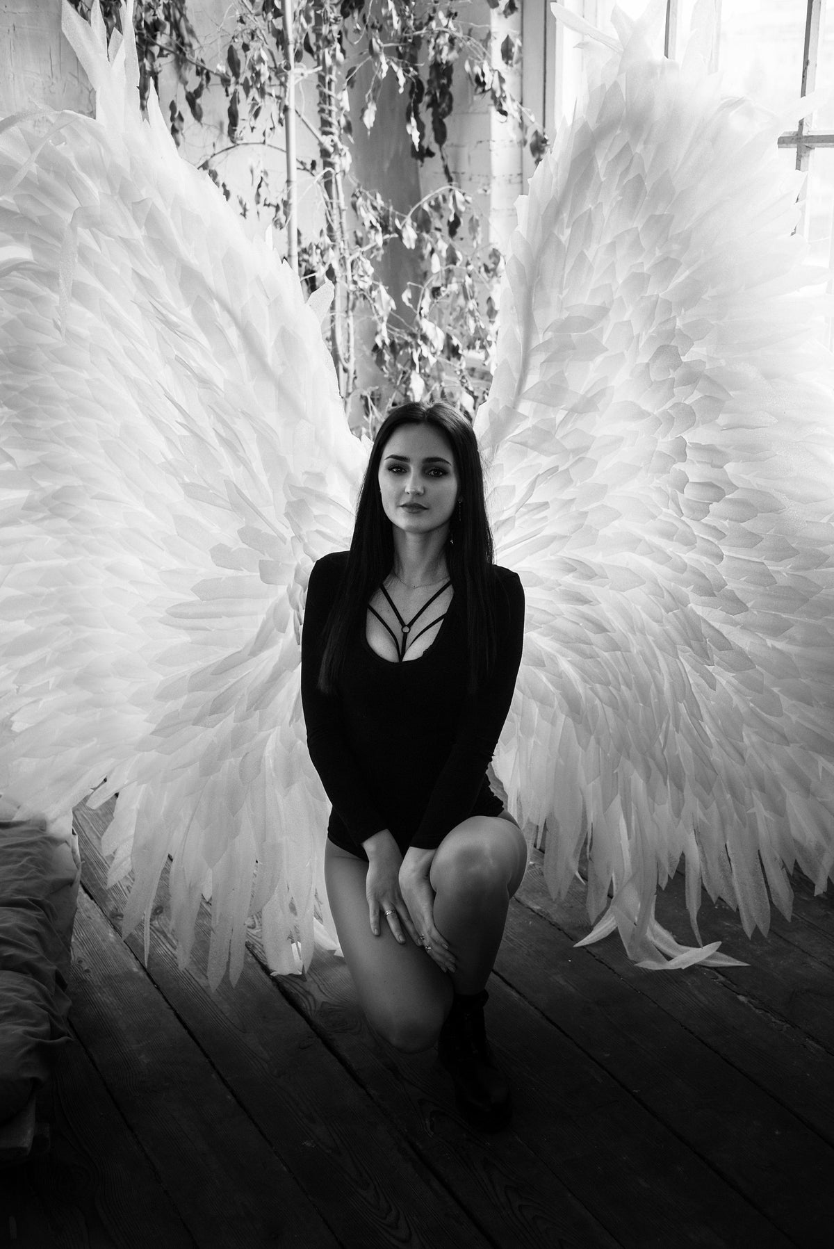 May include: A woman wearing a black long-sleeved top and white angel wings poses for a photo. She is kneeling on a wooden floor.