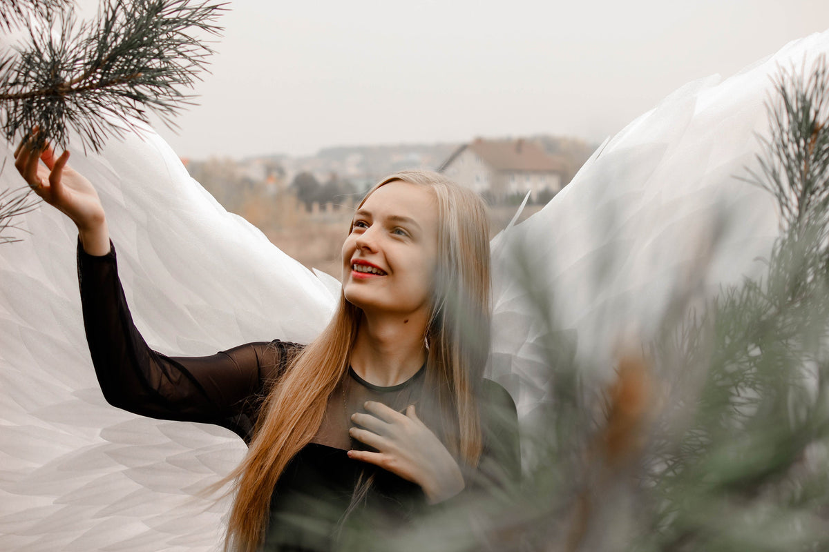 May include: A woman with long blonde hair and a black top, wearing large white angel wings. She is smiling and reaching towards a pine branch. The background shows a blurred landscape with buildings and trees.