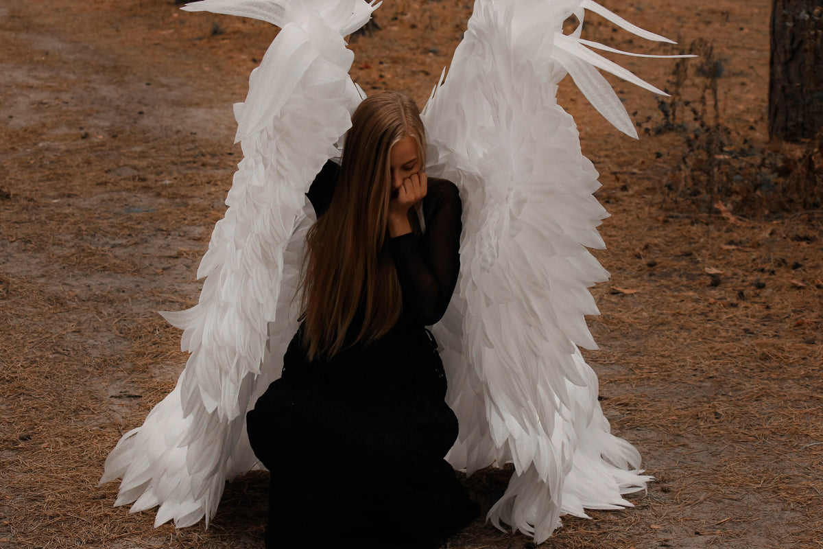 May include: A person wearing a black dress and large white angel wings, kneeling in a forest setting. The wings are detailed with layered feathers. The person's long, light brown hair cascades down, and they are covering their face.