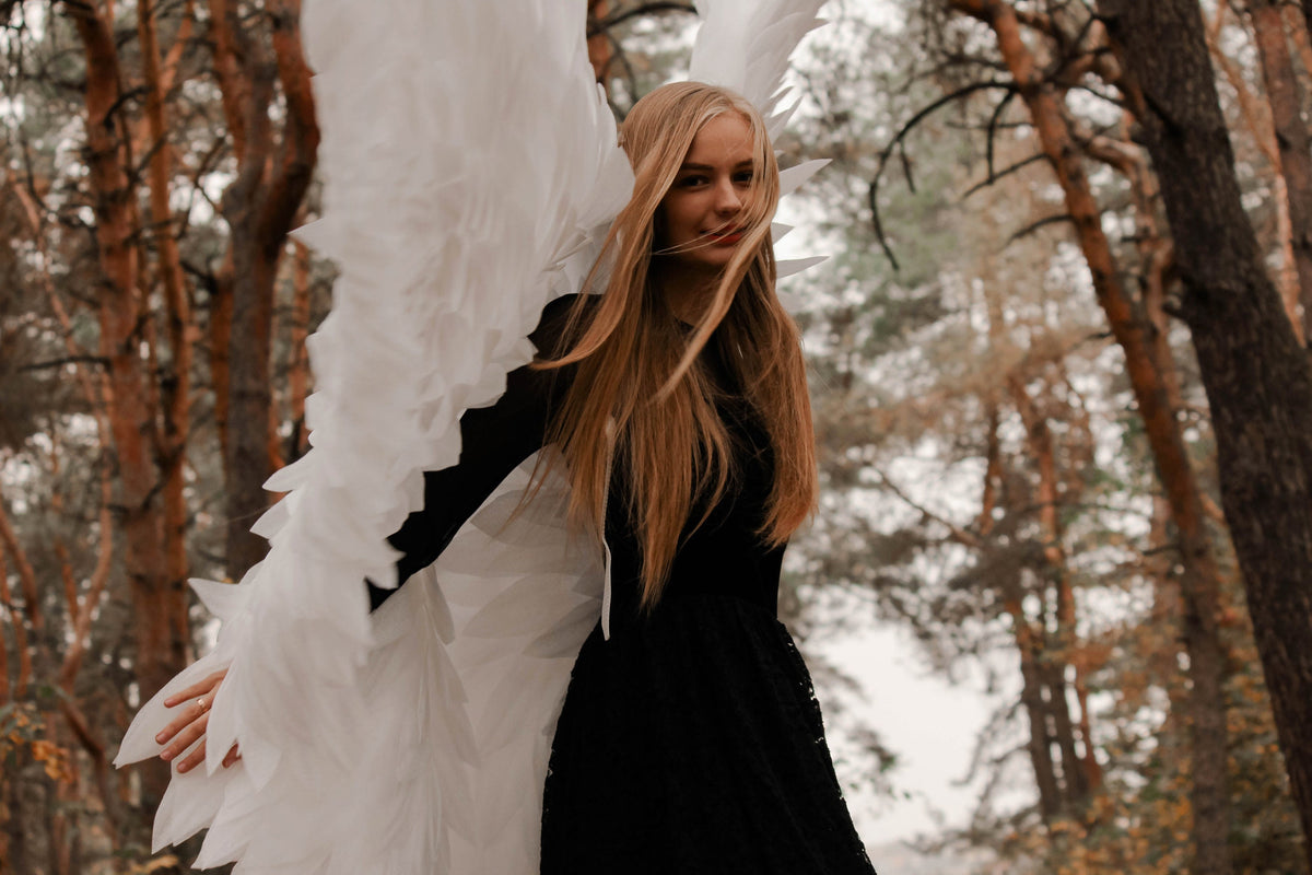 May include: A woman with long blonde hair and a black dress poses outdoors, wearing large white angel wings. The wings are made of layered, textured feathers. The background features a forest with tall trees and muted colors.