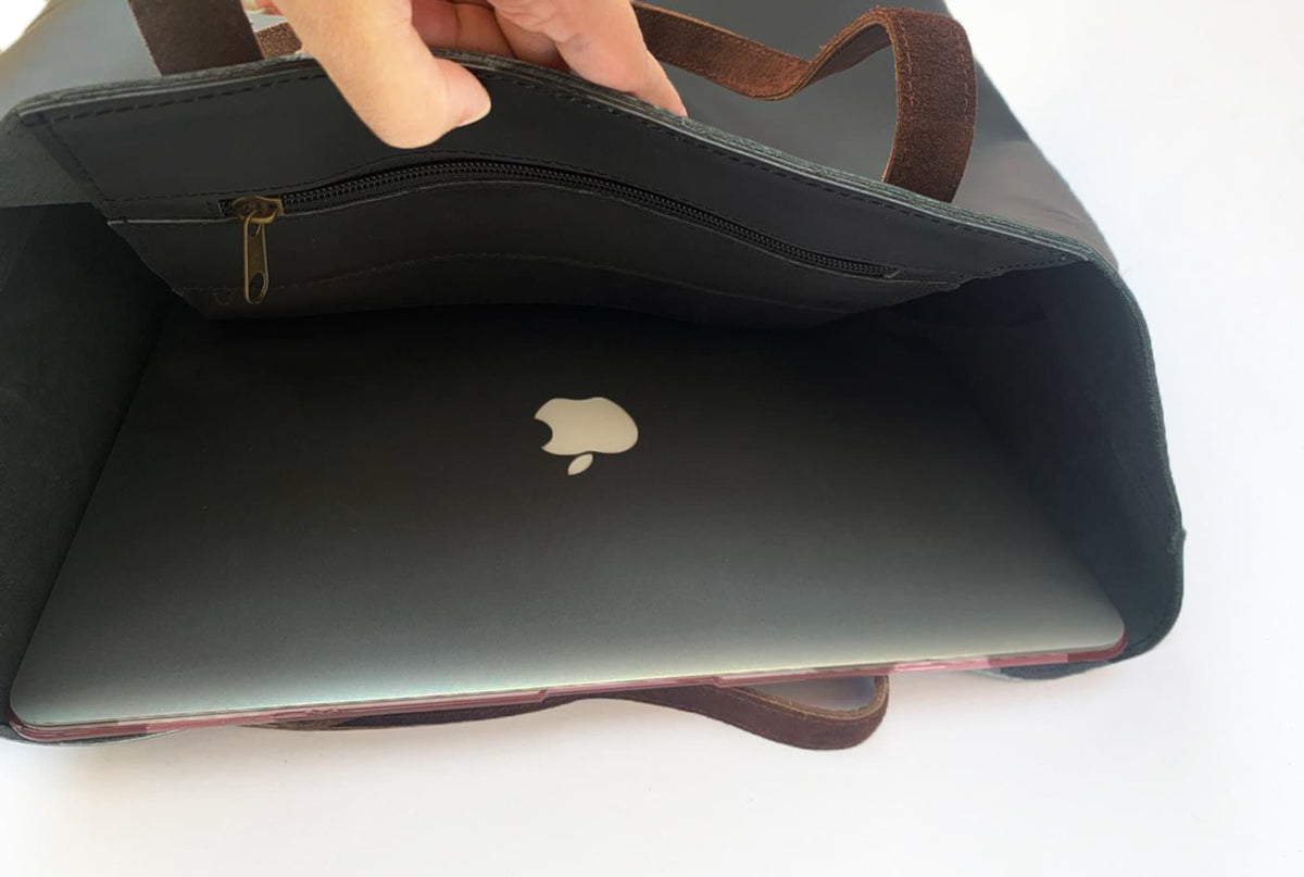 May include: Black leather tote bag with brown suede handles. The interior features a zippered pocket and holds a laptop with the Apple logo visible. The bag is open, revealing the contents.