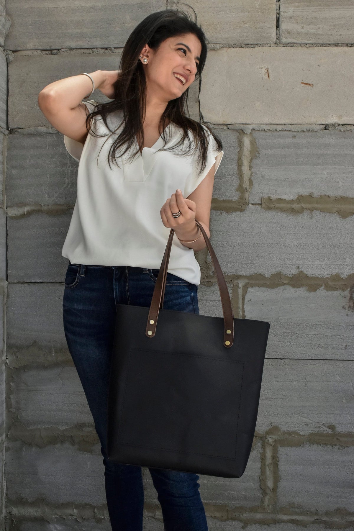 May include: A woman is holding a black leather tote bag with brown leather straps and gold accents. The bag has a large pocket on the front.