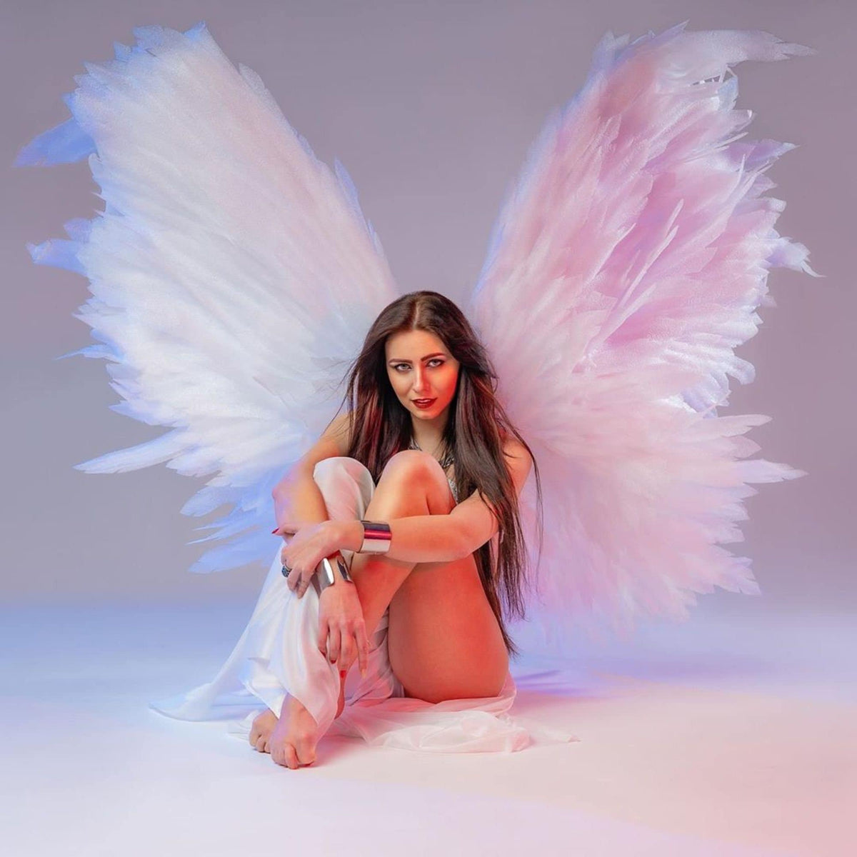 May include: A woman with long dark hair sits with her legs crossed, wearing a white flowing garment. She is wearing a silver bracelet on her wrist. She is surrounded by large white and pink angel wings.