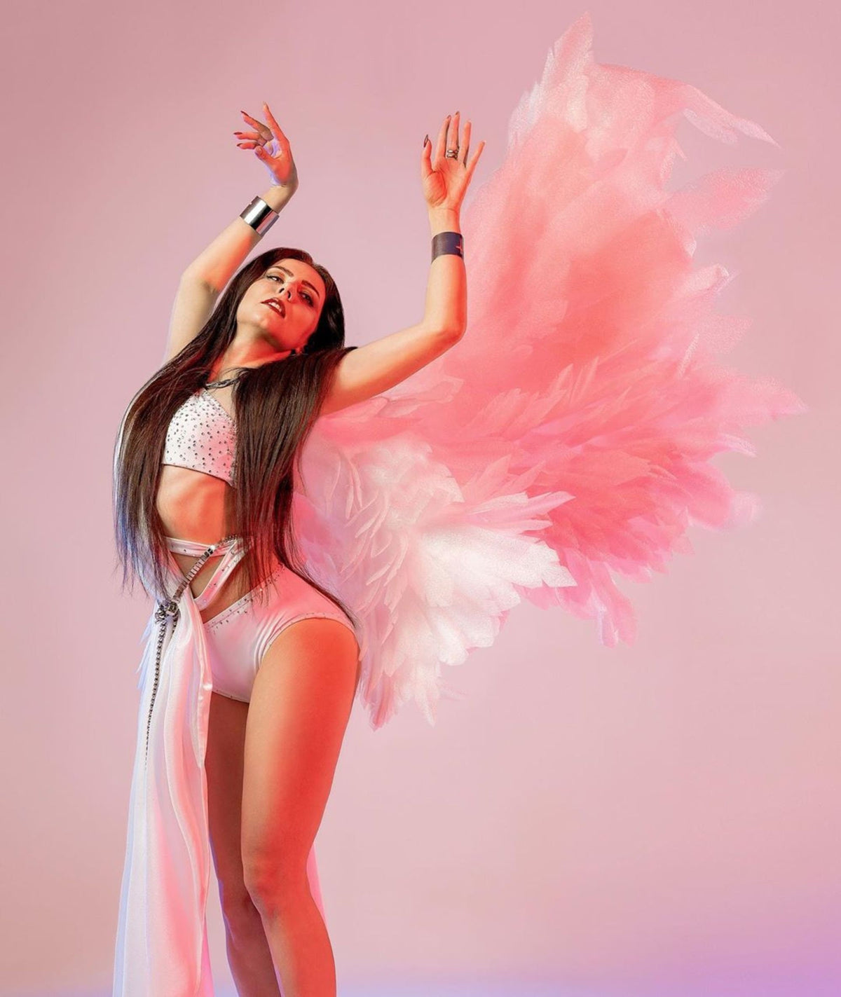 May include: A woman wearing a white costume with a pink feathered angel wing. The costume features a white top with rhinestones, a white skirt, and a white sash. The woman is posing with her arms raised above her head.
