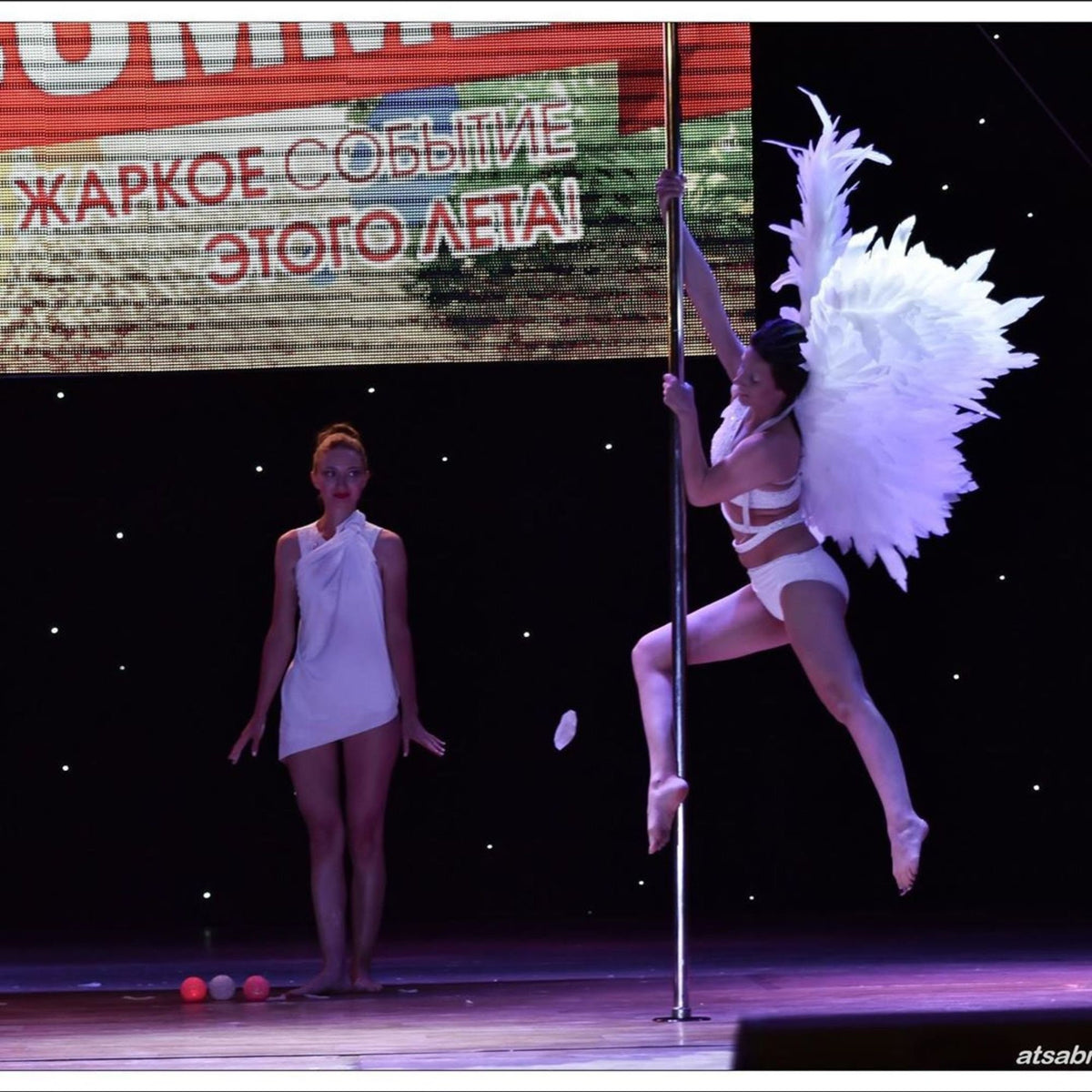 May include: A woman in a white outfit with angel wings performs a pole dance on a silver pole. A second woman in a white dress stands on the stage watching the performance. The background is a dark stage with a large screen displaying the text "ЖАРКОЕ СОБЫТИЕ ЭТОГО ЛЕТА!" in Russian.