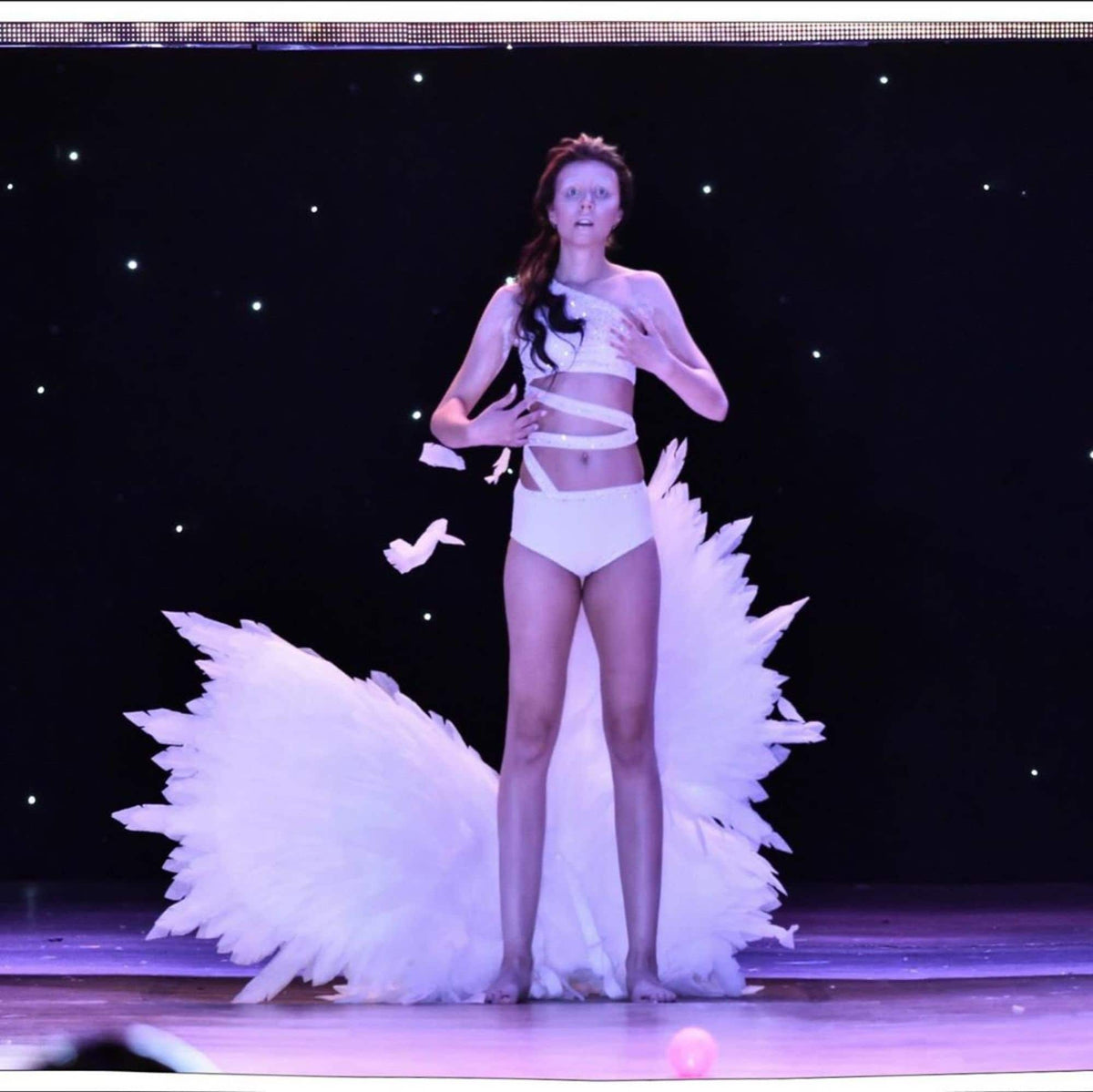 May include: A woman in a white costume with a large white feathered skirt and wings. She is standing on a stage with a dark background.