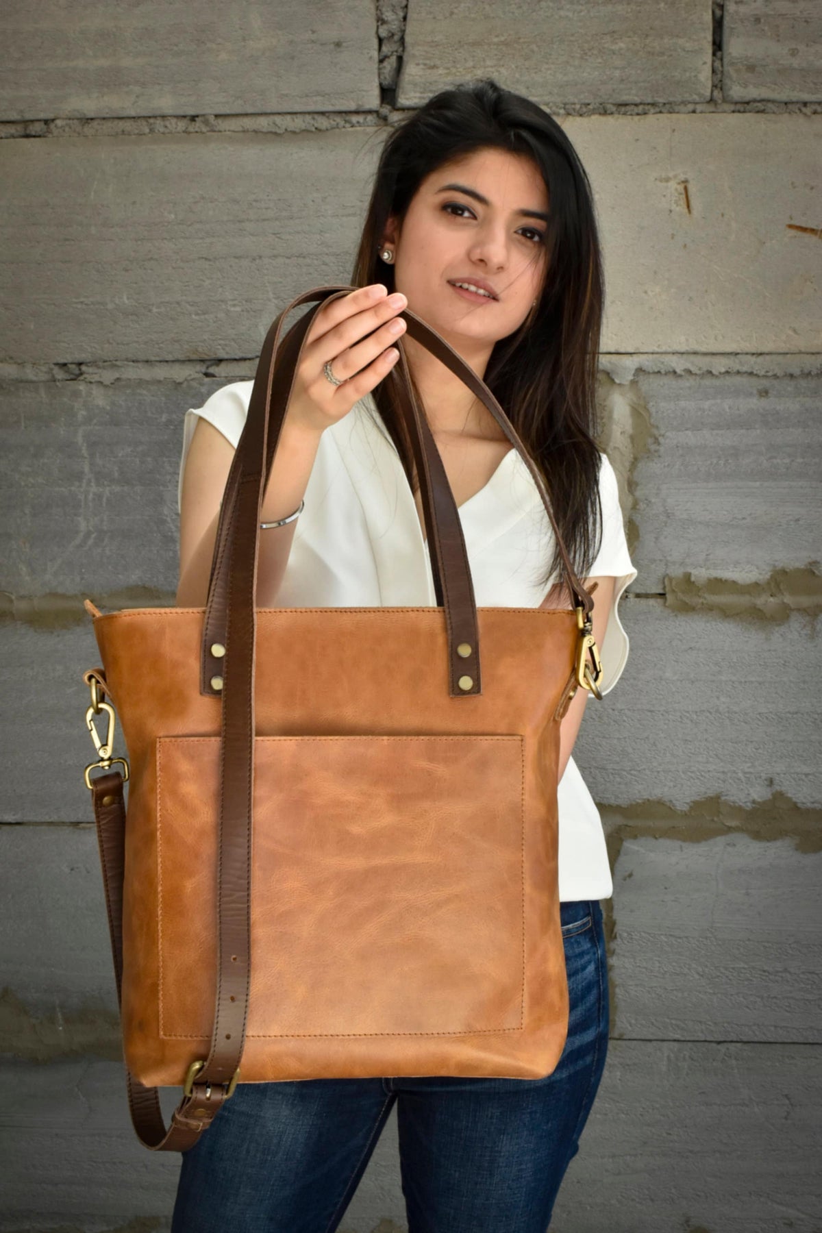 May include: A brown leather tote bag with a brown shoulder strap and a front pocket. The bag has gold metal hardware and is held by a person wearing a white shirt and blue jeans.