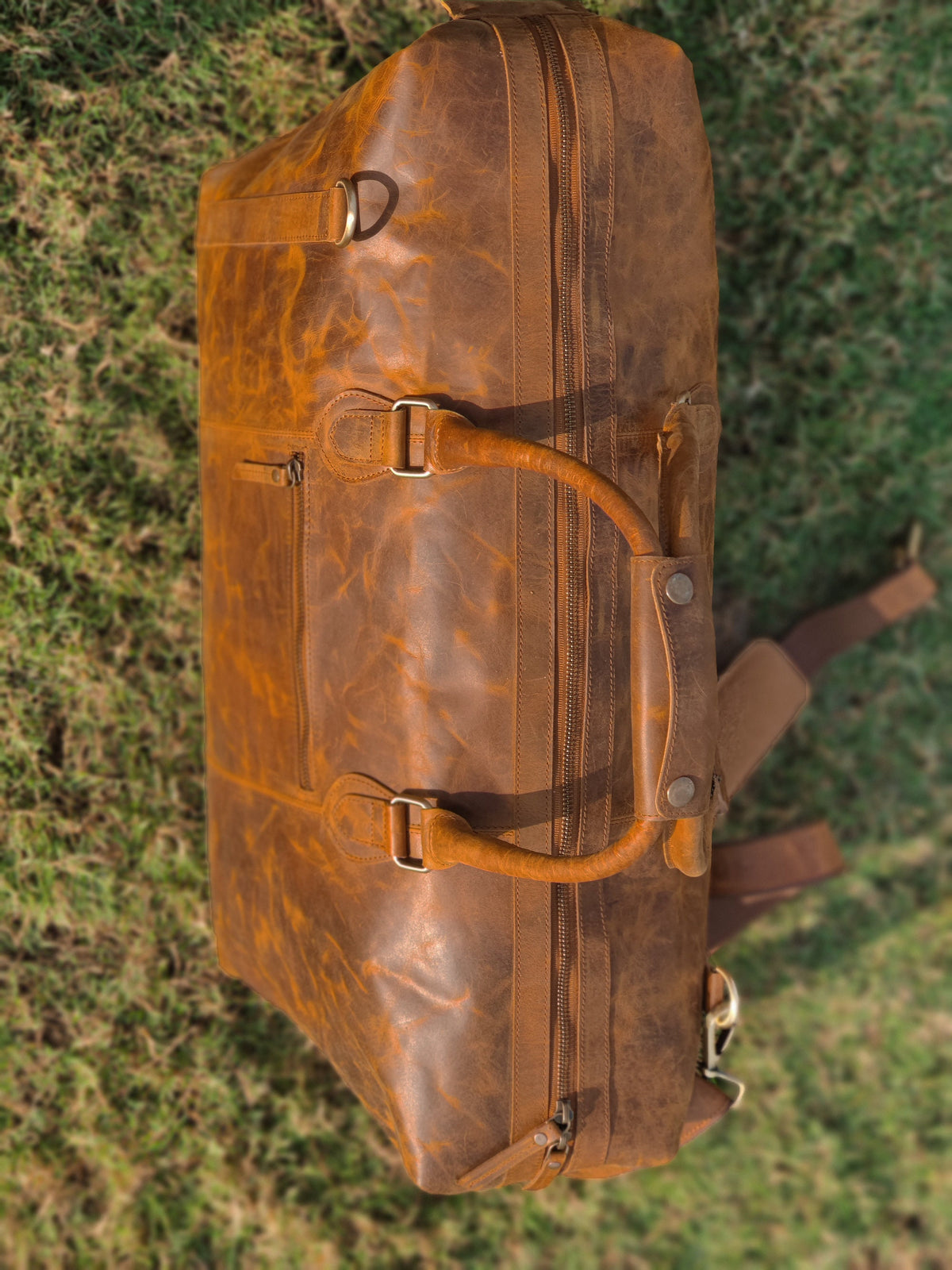 May include: A brown leather duffel bag with multiple zippered compartments and a top handle. The bag has a distressed look and is made of a thick, durable leather.
