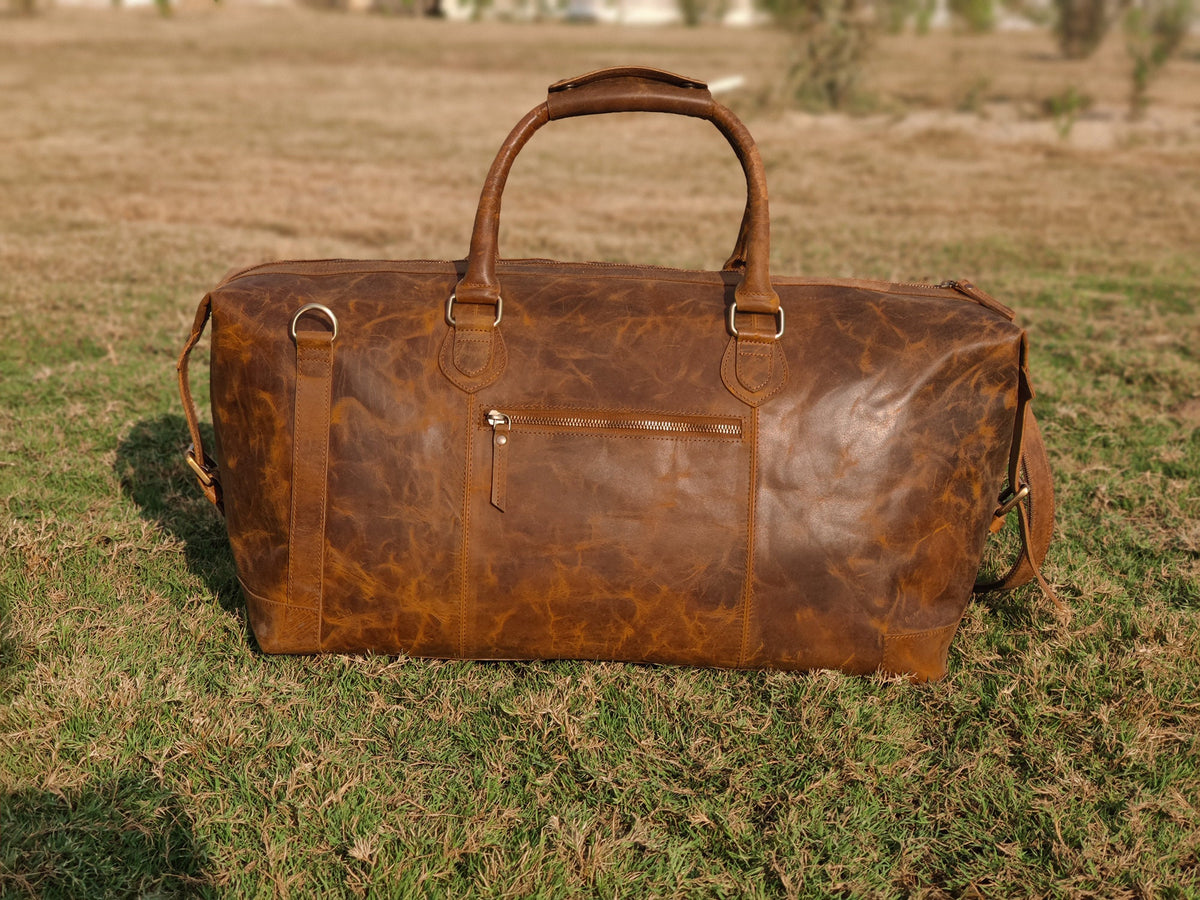 May include: A brown leather duffel bag with a top handle and a zippered pocket on the front. The bag has a distressed look and is made of high-quality leather.
