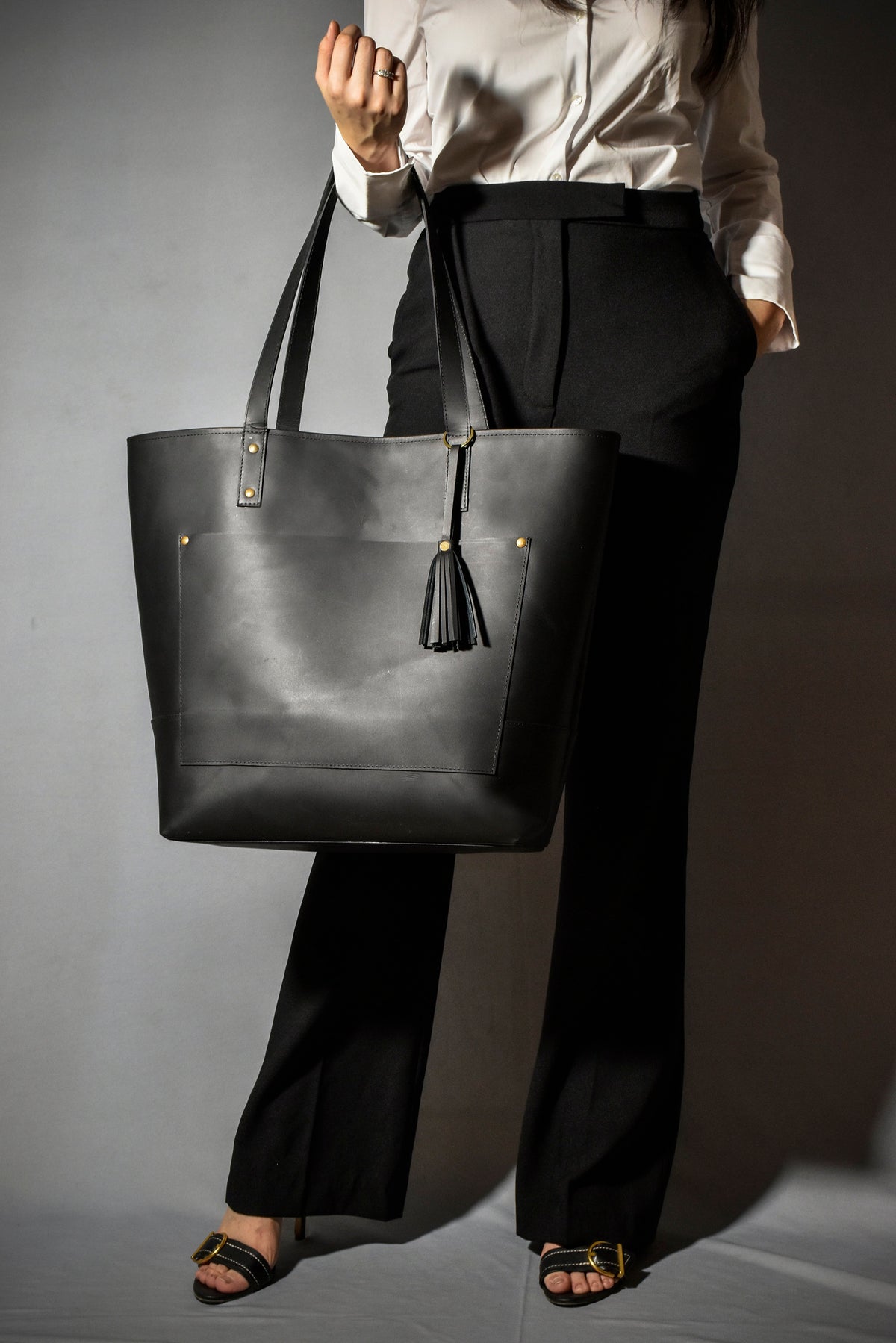 May include: A black leather tote bag with a gold tassel and a front pocket. The bag has a top handle and two shoulder straps.