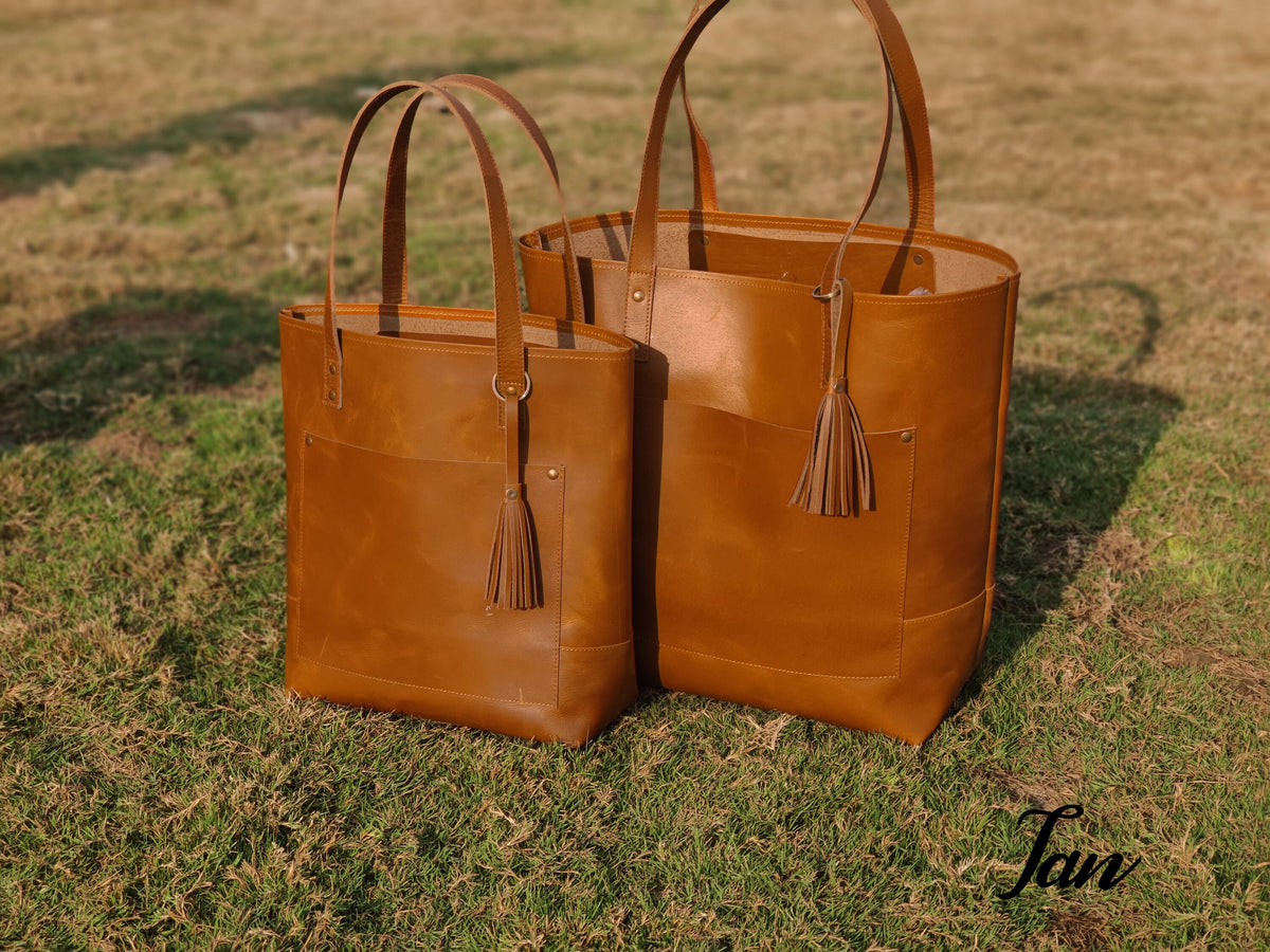 May include: Two brown leather tote bags with a tassel detail. The larger bag is behind the smaller bag.
