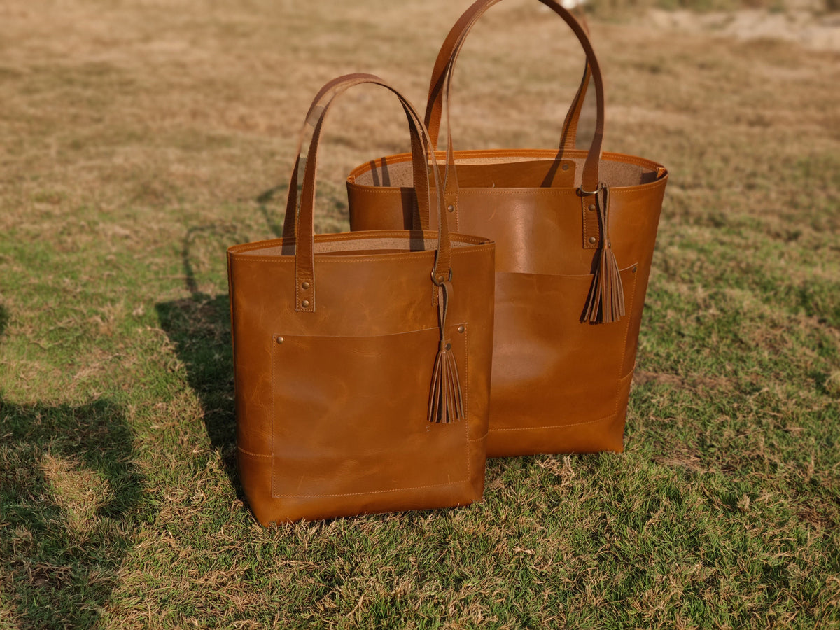May include: Two brown leather tote bags with tassels. The larger bag is behind the smaller bag.