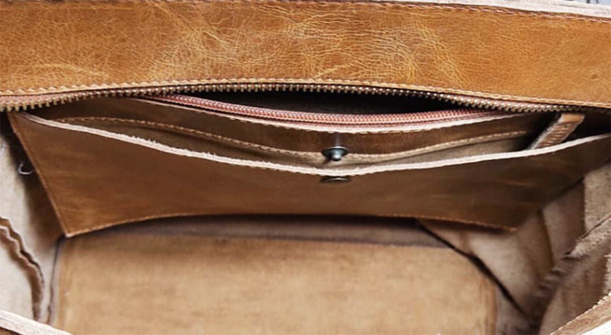 May include: Close-up of a tan leather bag interior. The bag features multiple compartments, including a zippered pocket and several open pockets. The leather has a distressed appearance, with visible texture and variations in color. A metal snap secures one of the pockets.