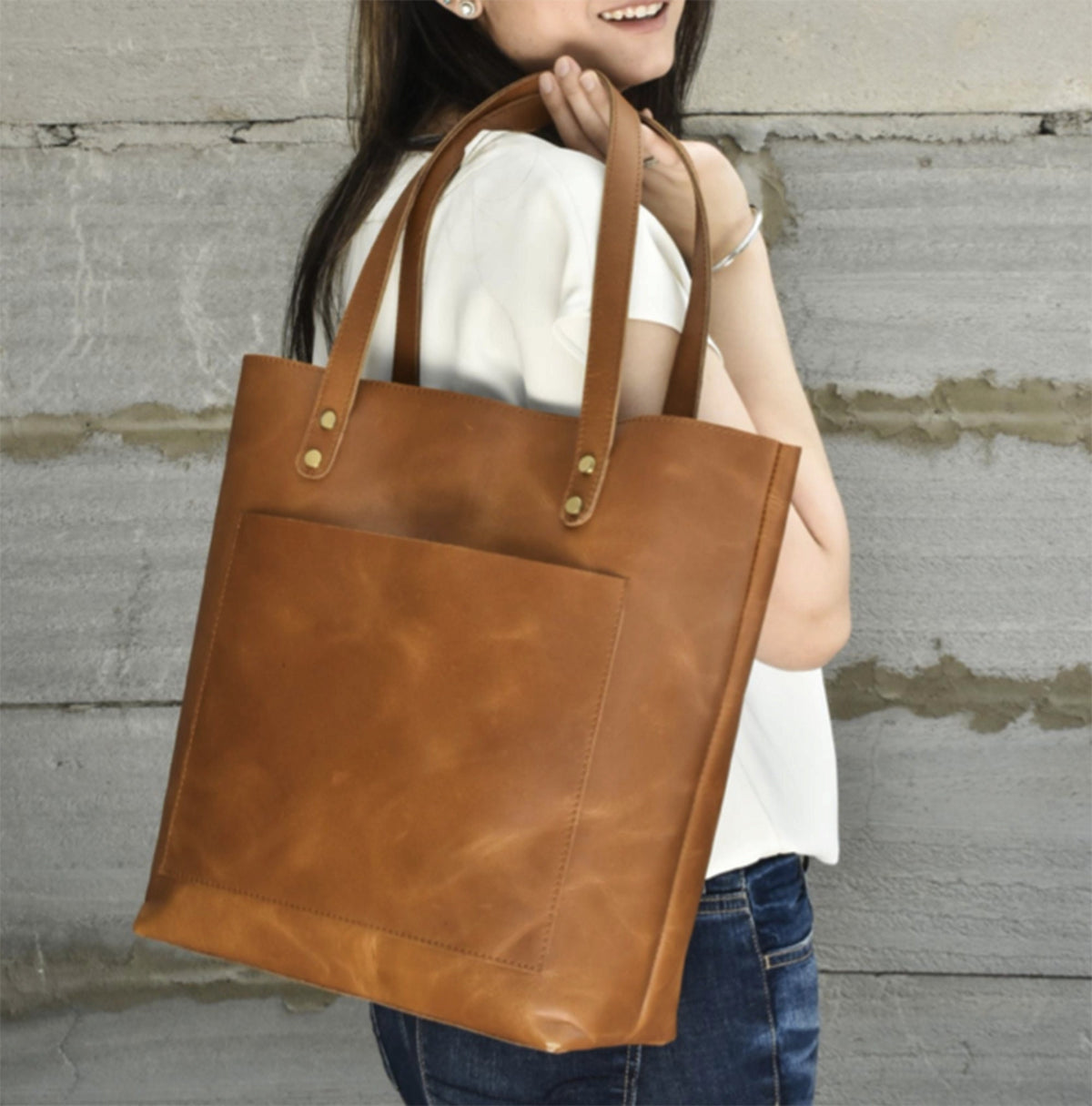 May include: A brown leather tote bag with a front pocket and two handles. The bag is a light brown color and has brass rivets at the top of the handles. The bag is being held by a person wearing a white shirt and blue jeans.