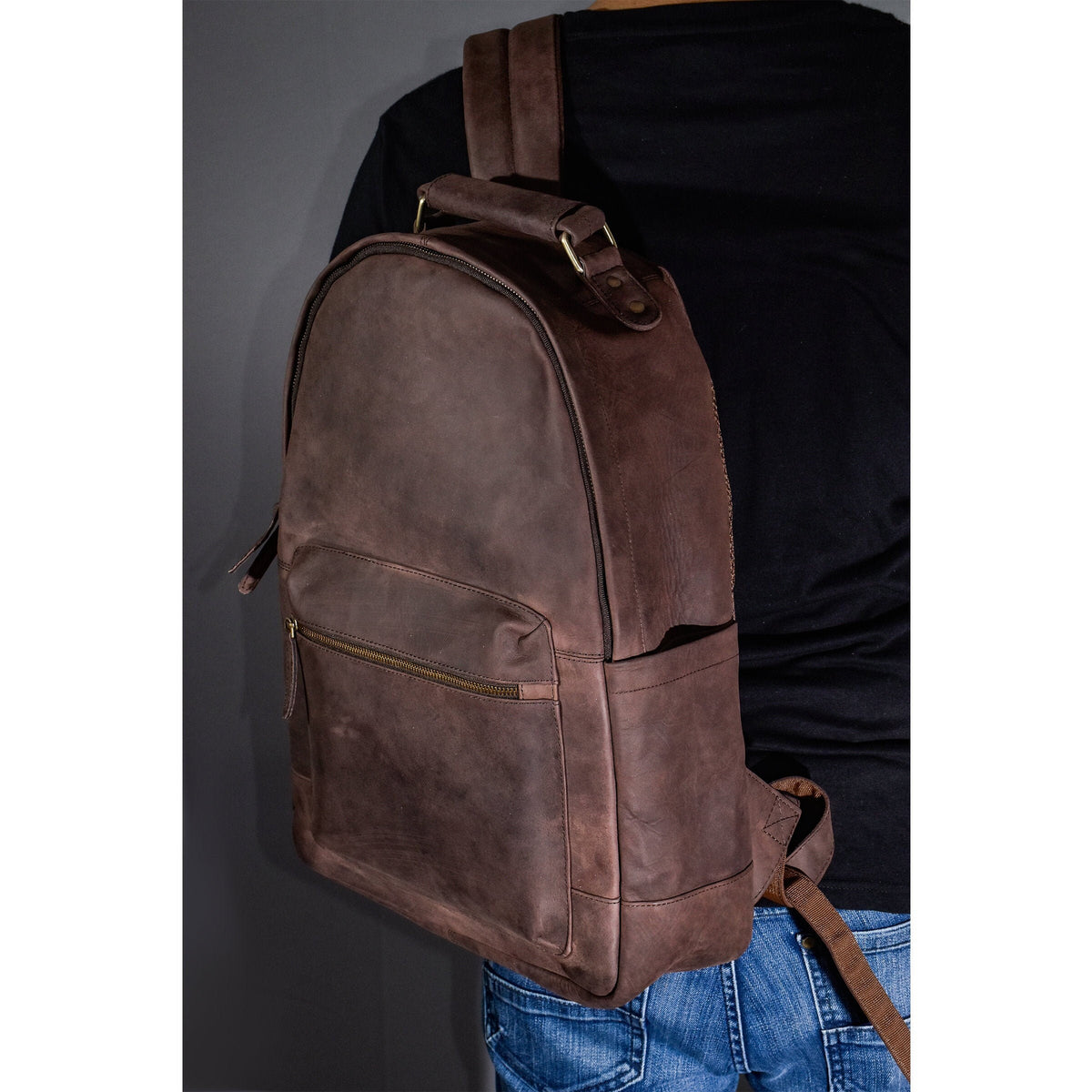 May include: A brown leather backpack with a top handle and a zippered pocket on the front. The backpack is being worn by a person.