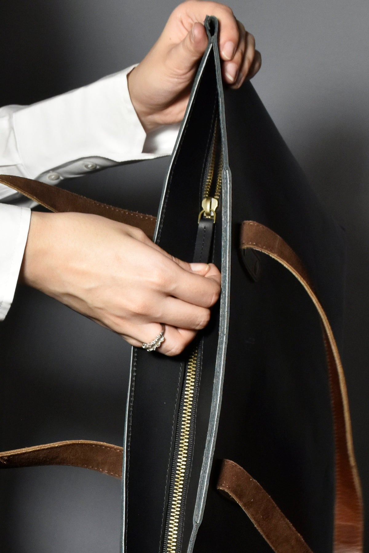 May include: A black leather tote bag with brown leather straps and a gold zipper. The bag is open and the zipper is pulled down.