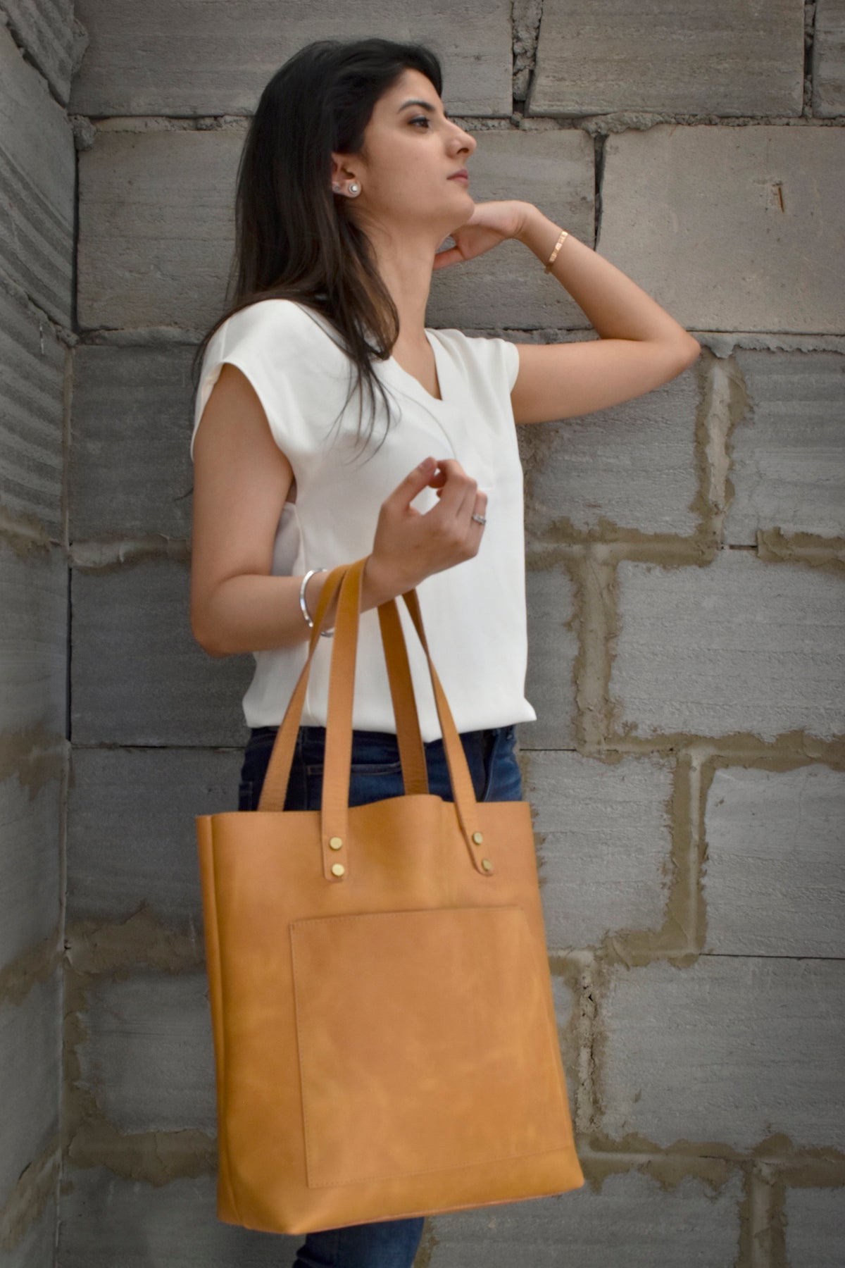 May include: A tan leather tote bag with a rectangular shape and a front pocket. The bag has two handles and is held by a person wearing a white top and blue jeans. The bag is against a gray brick wall.