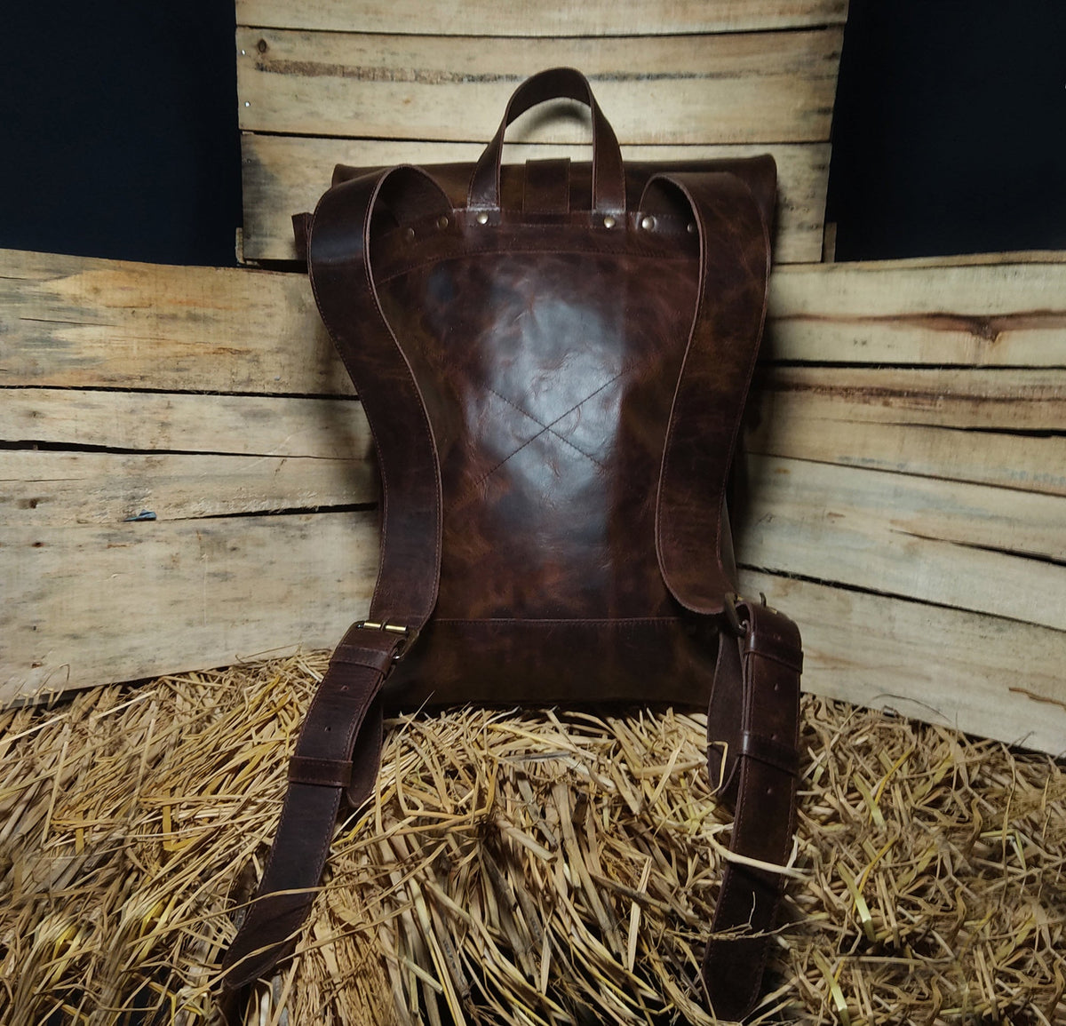May include: A dark brown leather backpack featuring wide adjustable shoulder straps, a top carry handle, and metal rivets securing the straps to the main body. The back panel displays a subtle stitched X pattern across the center surface.
