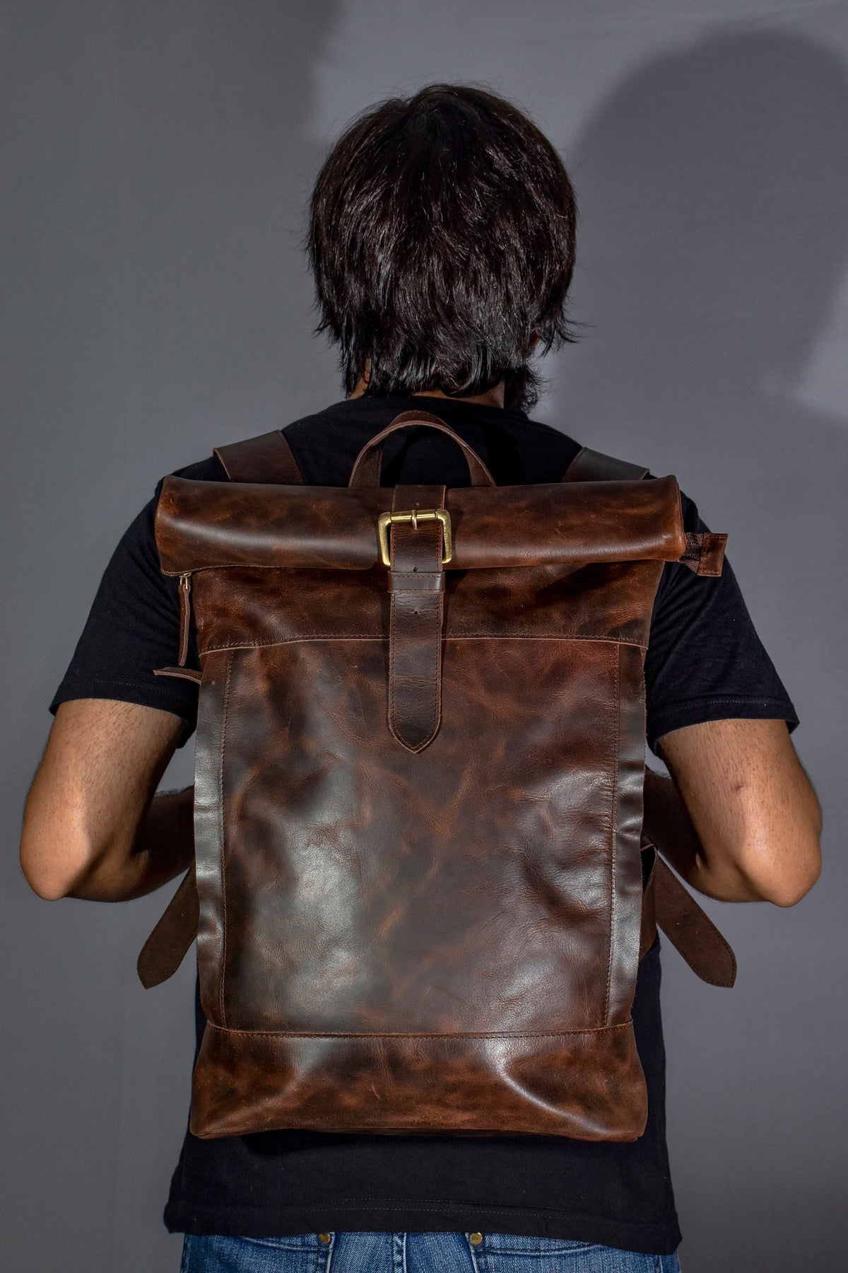 May include: A brown leather backpack with a roll-top closure and a buckle. The backpack has a distressed look and is worn by a person.
