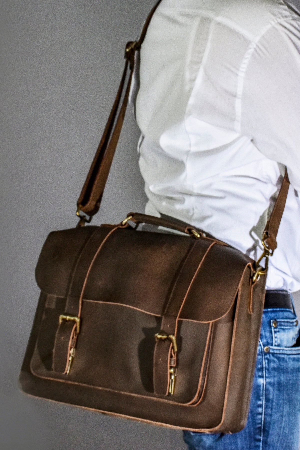 May include: A brown leather messenger bag with a shoulder strap and a top handle. The bag has two front buckles and a flap closure. The bag is worn over the shoulder.