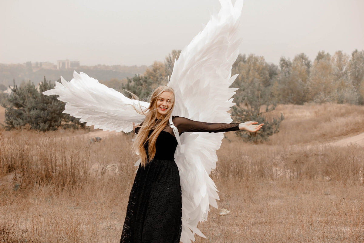 May include: A woman with long blonde hair and a black dress poses outdoors with large white angel wings. The wings are made of feathers and are spread wide, creating a dramatic effect against the natural background.