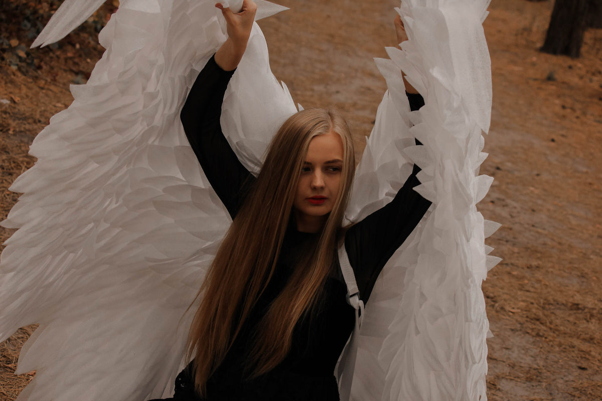 May include: A person wearing a black top and holding large white angel wings. The wings are made of layered, textured material. The background is a blurred outdoor scene with brown and tan tones.