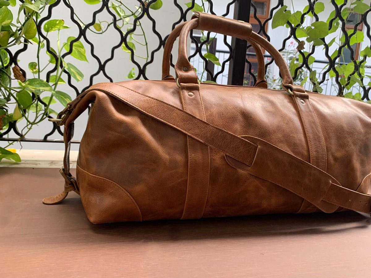 May include: A brown leather duffel bag with a top handle and a shoulder strap. The bag has a distressed look and is made of high-quality leather.
