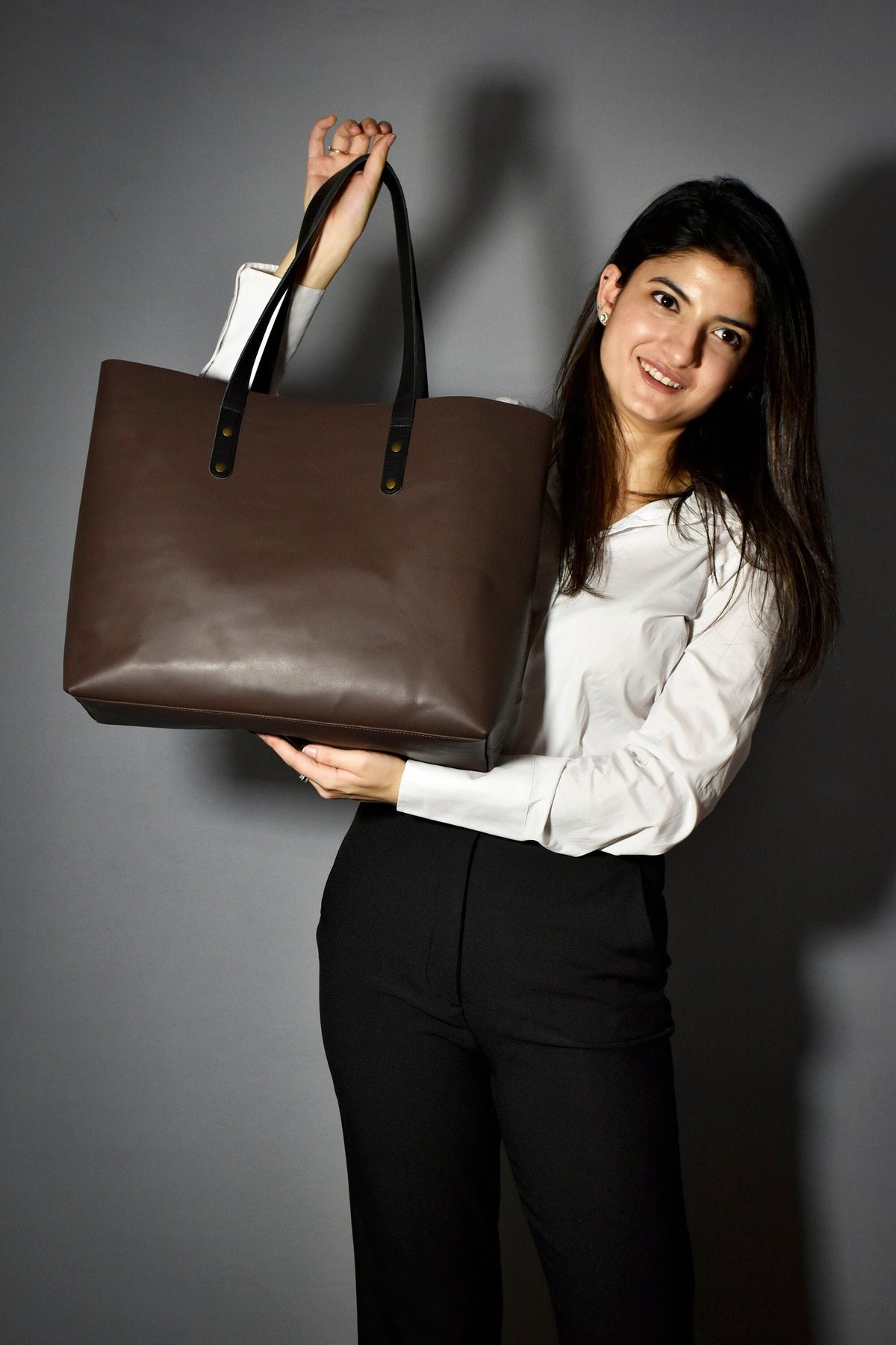 May include: A brown leather tote bag with black handles and gold accents. The bag is being held by a person wearing a white shirt and black pants.