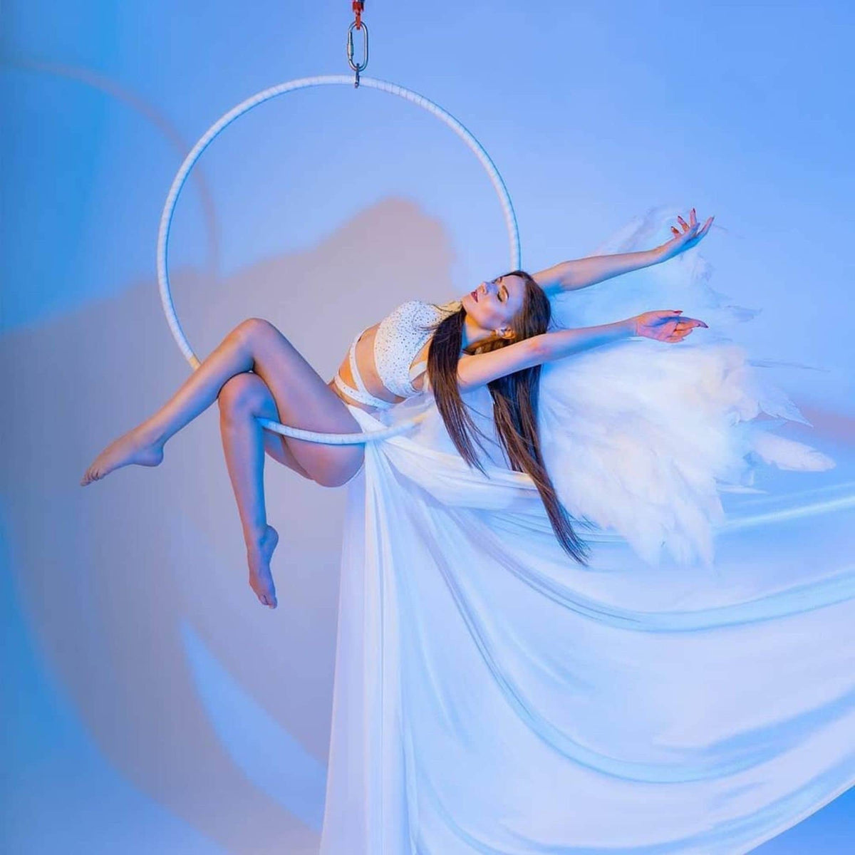 May include: A woman in a white leotard and white wings is performing aerial acrobatics in a white hoop suspended from the ceiling. The background is a light blue.
