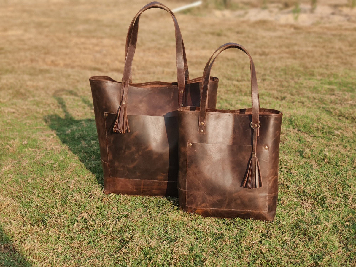 May include: Two brown leather tote bags with a tassel detail. The larger bag has a front pocket. Both bags have a distressed leather look.