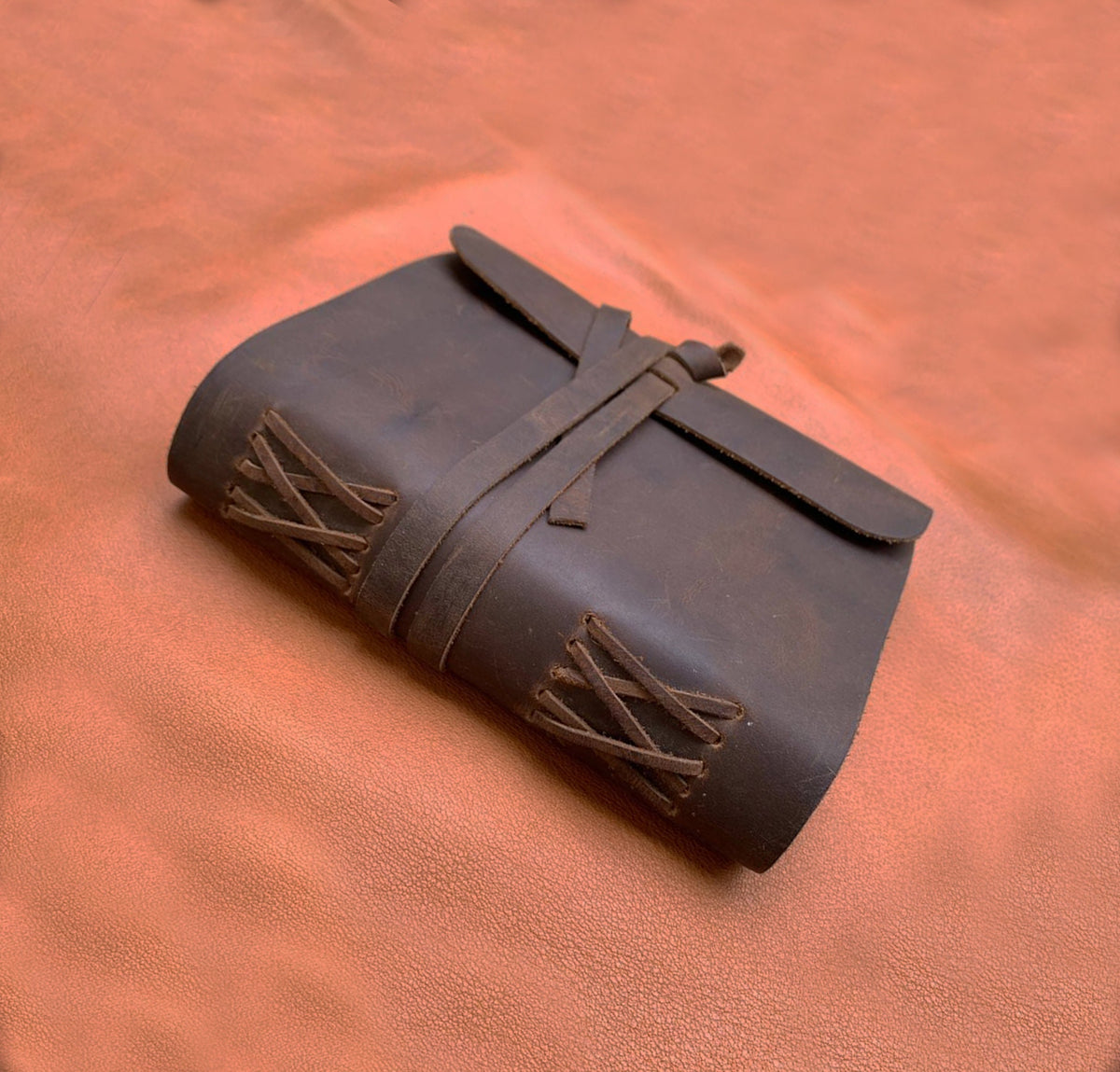 May include: A brown leather journal with a strap closure and decorative stitching. The journal is closed and lying on a brown leather surface.