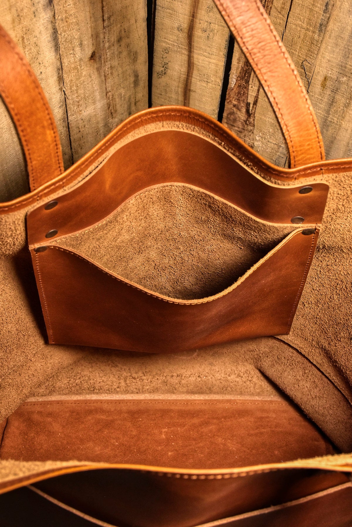 May include: A brown leather tote bag with two handles and an interior pocket. The bag has a natural, rustic appearance with visible stitching and a textured surface. The interior is a lighter shade of brown, and the bag appears to be handmade.