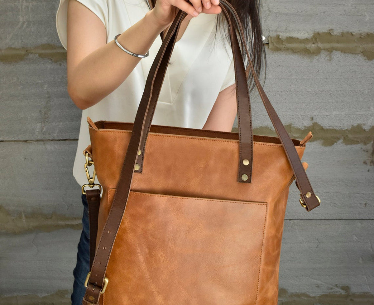 May include: A tan leather tote bag featuring a large exterior pocket, dark brown leather handles, and an adjustable dark brown shoulder strap attached with brass-toned metal hardware, including rivets and swivel clasps on the sides.