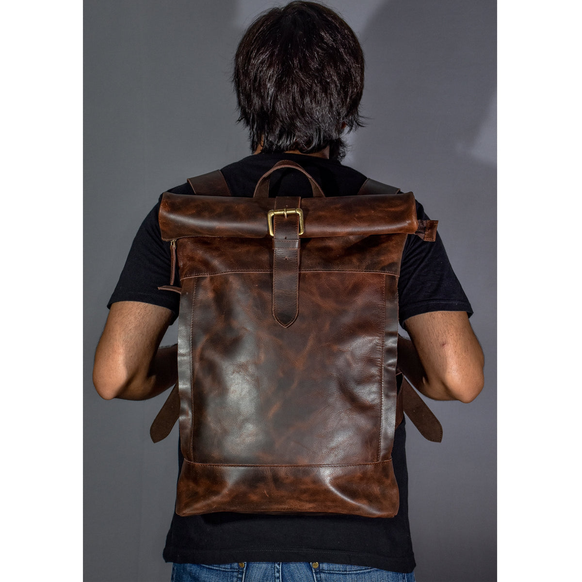 May include: A brown leather backpack with a roll-top closure and a brass buckle. The backpack has a distressed look and is worn by a person.