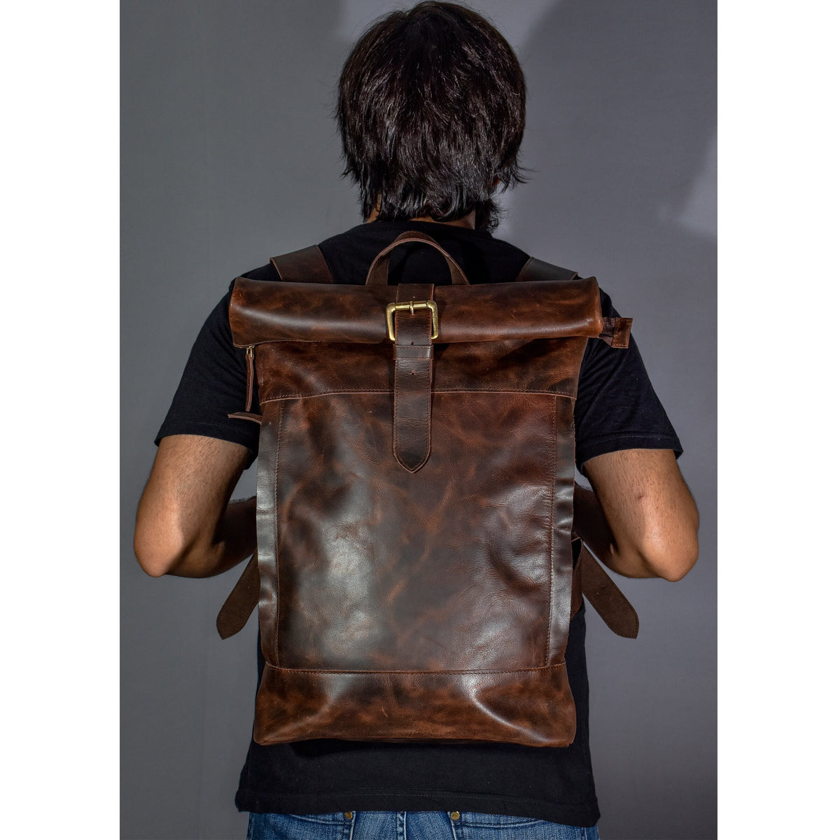 May include: A brown leather backpack with a roll-top closure and a brass buckle. The backpack has a distressed look and is worn by a person.